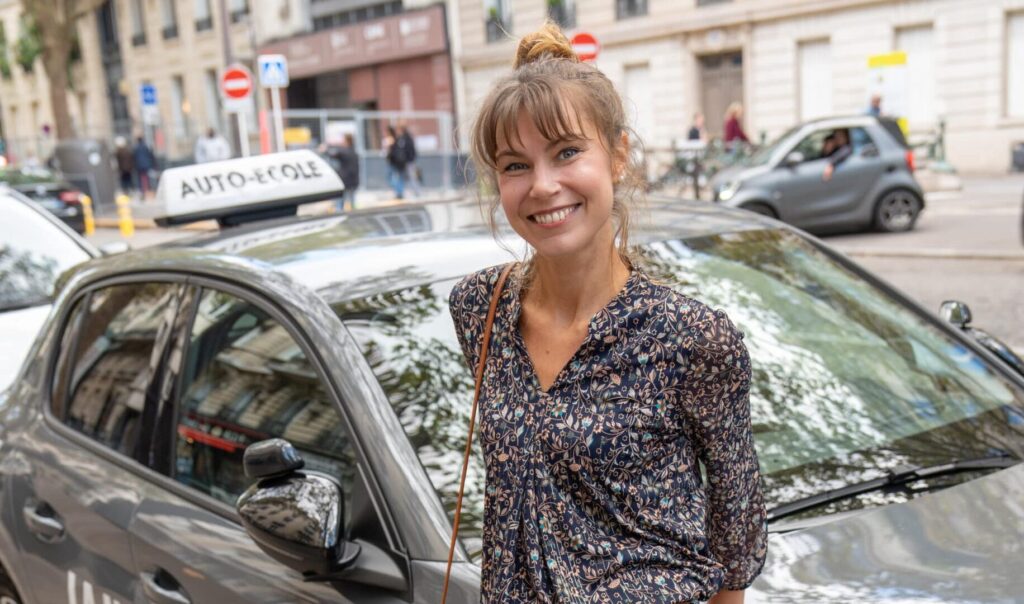 Jeune femme souriante debout devant une voiture d’auto-école en ville, avec un panneau “auto-école” sur le toit du véhicule.