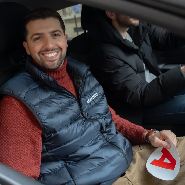 un homme souriant assis à l'avant d'une voiture côté passager, tient à la main le macaron A jeune conducteur