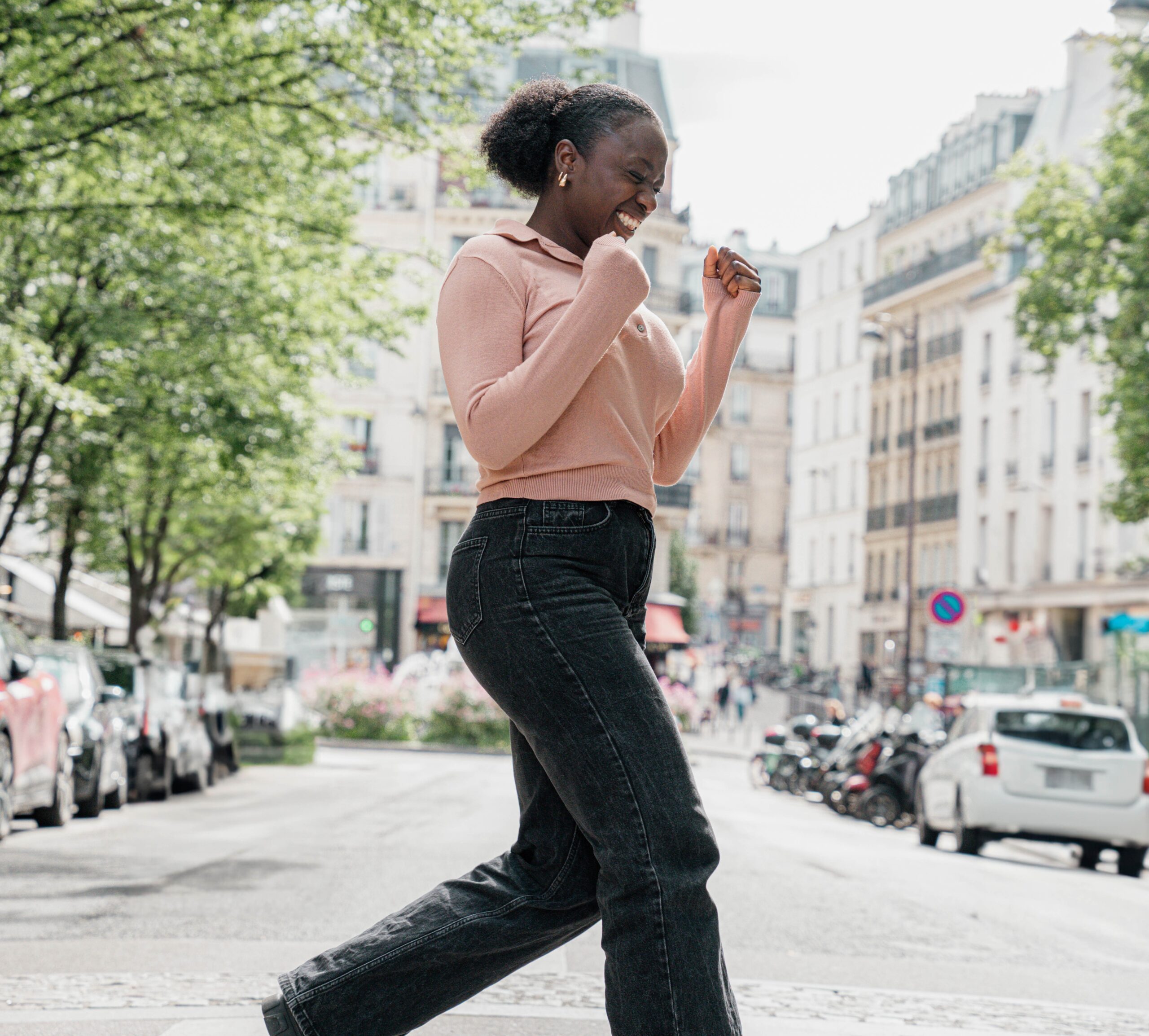 une jeune femme, pull rose, traverse la rue. Elle est très heureuse
